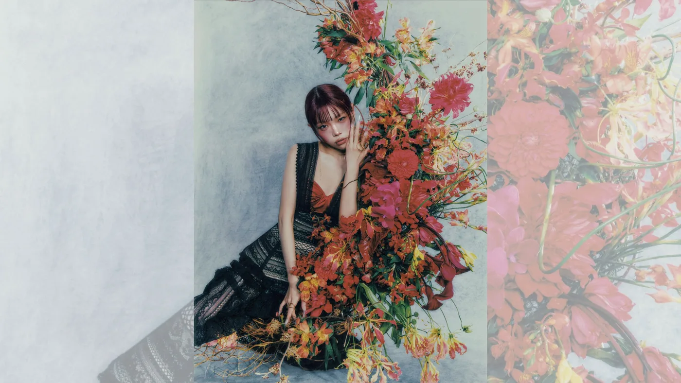 AiNA THE END sentada junto a un gran arreglo de flores rojas y naranjas, con vestido negro de encaje, sobre un fondo gris azulado