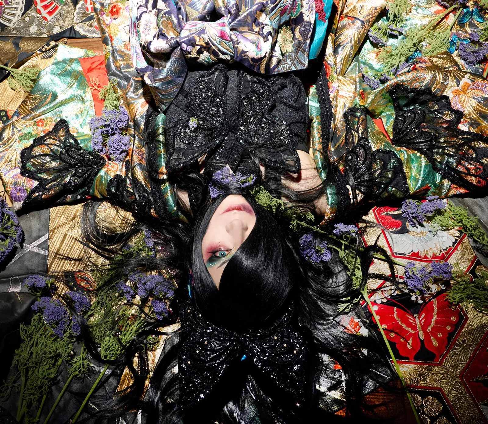 Foto promocional de ASAGI: recostado boca arriba con el cabello negro sobre la cara, vestido de negro sobre kimonos y obis bordados de colores, rodeado de flores moradas y ramas verdes