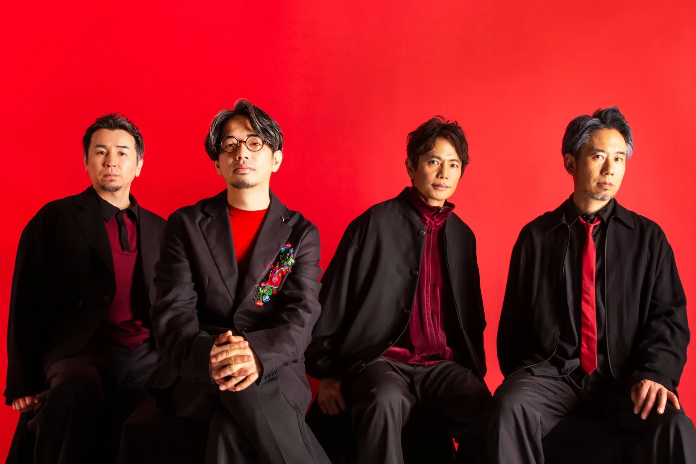 Foto oficial de los cuatro integrantes de ASIAN KUNG-FU GENERATION sentados en fila, vestidos de negro con camisas y corbatas rojas, frente a un fondo rojo