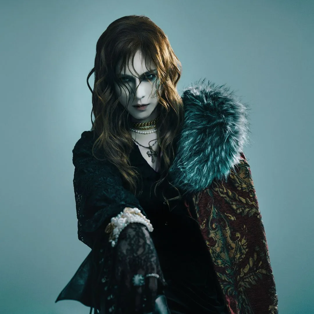 Foto promocional oficial de KAMIJO con abrigo de brocado y cuello de piel sobre fondo azul verdoso