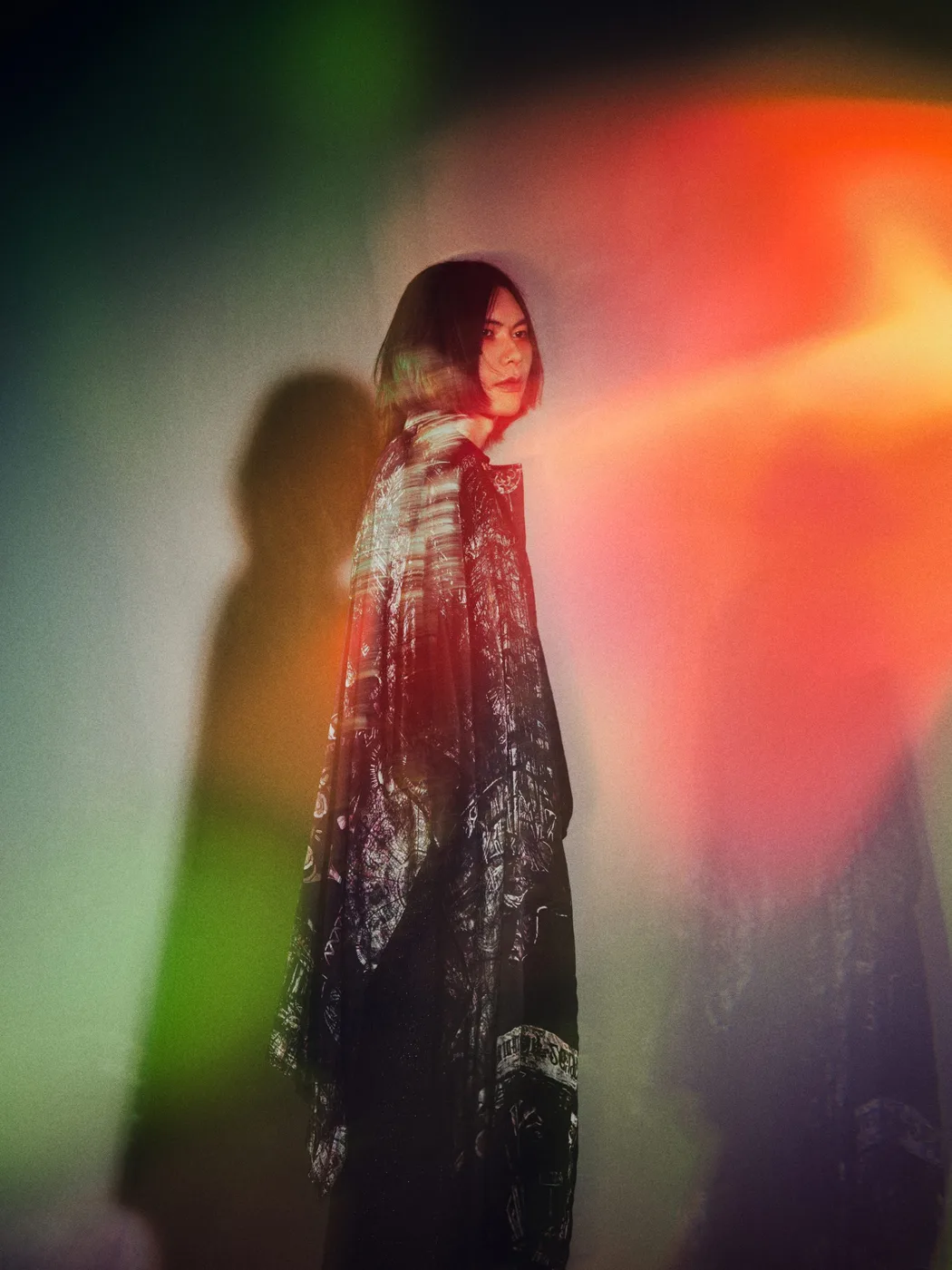 Foto oficial de Kenshi Yonezu con un abrigo largo estampado, frente a una pared iluminada con luces de colores